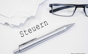 Grunderwerbsteuer: Steuervergünstigung nach § 6a des Grunderwerbsteuergesetzes das Wort "Steuern" steht auf weißem Grund neben einem Kugelschreiber, gestapeltem Papier und einer Brille